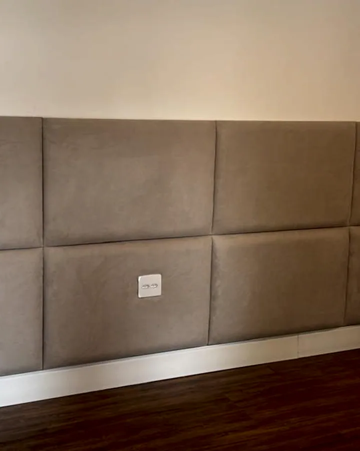 Painel de cabeceira modular em retângulos de suede fendi, instalado isoladamente em uma parede branca lisa.