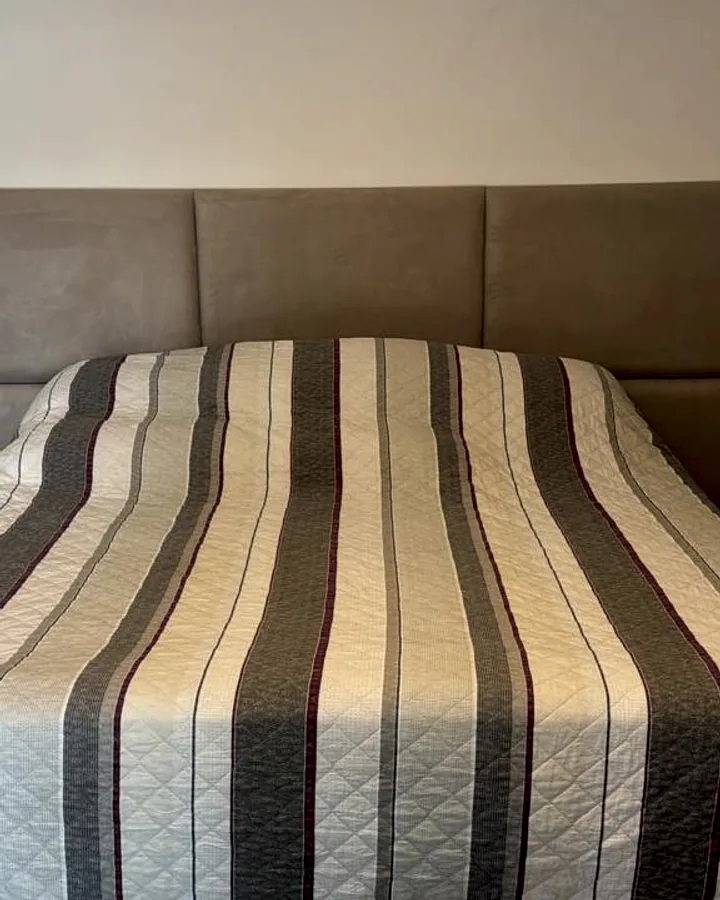 Cabeceira em painéis modulares horizontais na cor bege fendi, montada atrás de cama arrumada com colcha listrada.