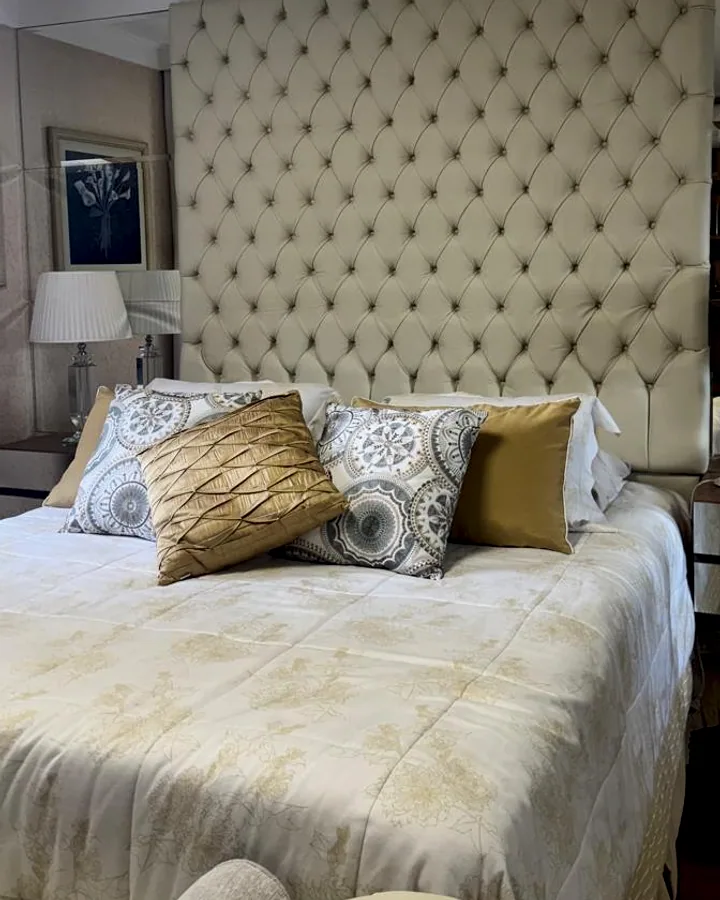 Cama com cabeceira alta estofada em capitonê bege claro, fixada sobre uma parede revestida de espelhos.