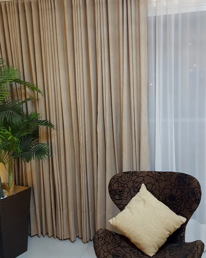Cortina bege em prega wave combinada com voil branco em ambiente interno com poltrona decorativa.