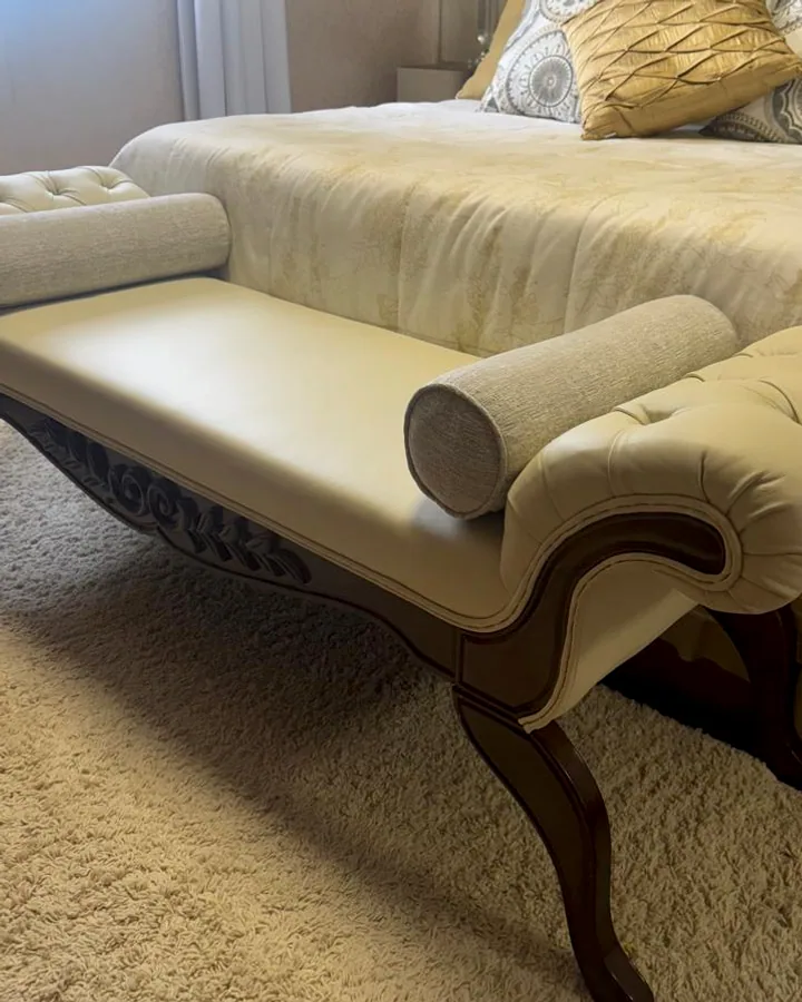 Recamier clássico em couro sintético bege com acabamento capitonê e detalhes em madeira posicionado ao pé de uma cama.