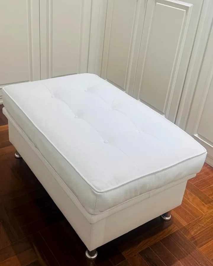 Puff retangular em couro sintético branco com acabamento capitonê posicionado em um piso de madeira em frente a armários brancos.