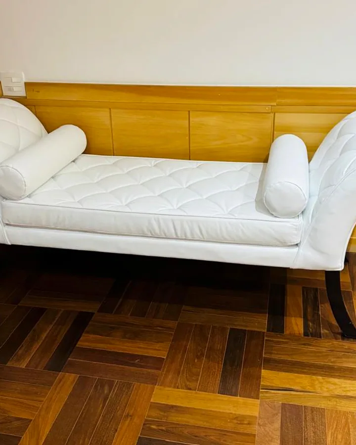 Recamier branco em capitonê com almofadas cilíndricas sobre um piso de taco de madeira.