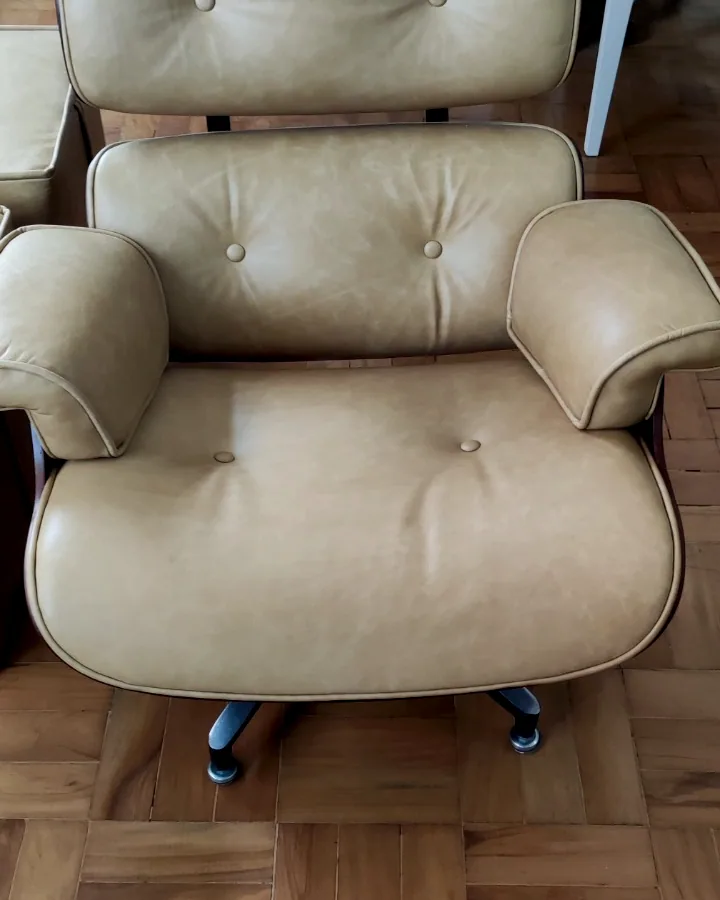 Poltrona Charles Eames estofada em couro bege com estrutura em madeira e base metálica sobre piso de taco de madeira.