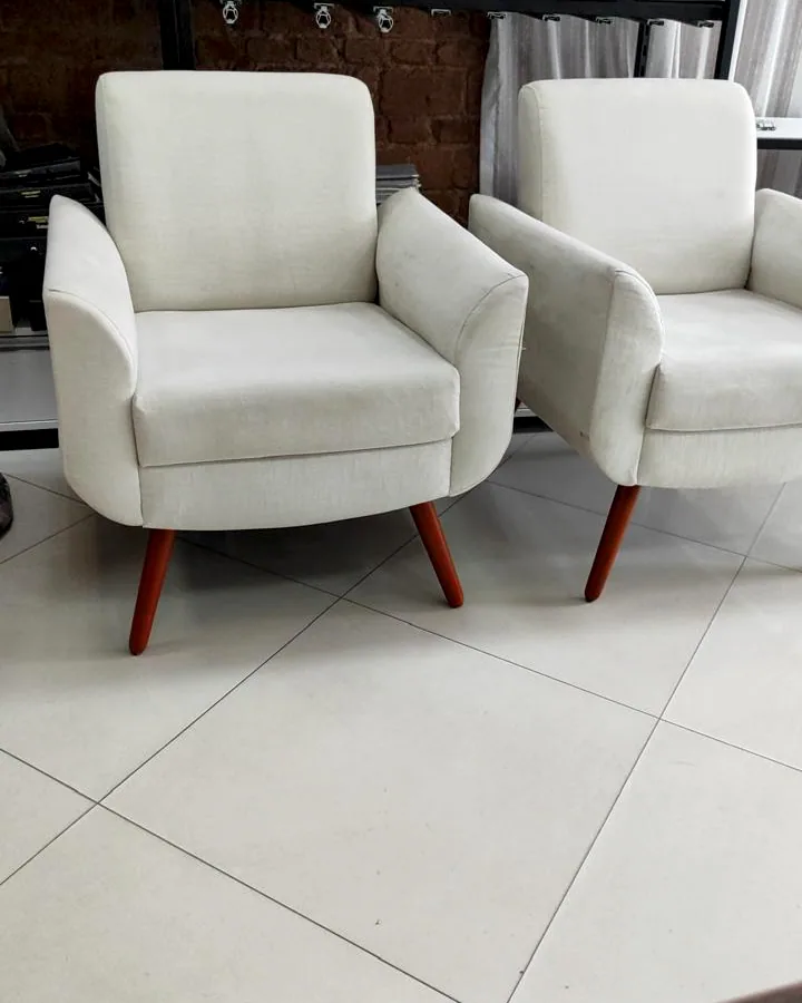 Par de poltronas idênticas com estofado em tecido claro e pés palito de madeira, em frente a estante com amostras.