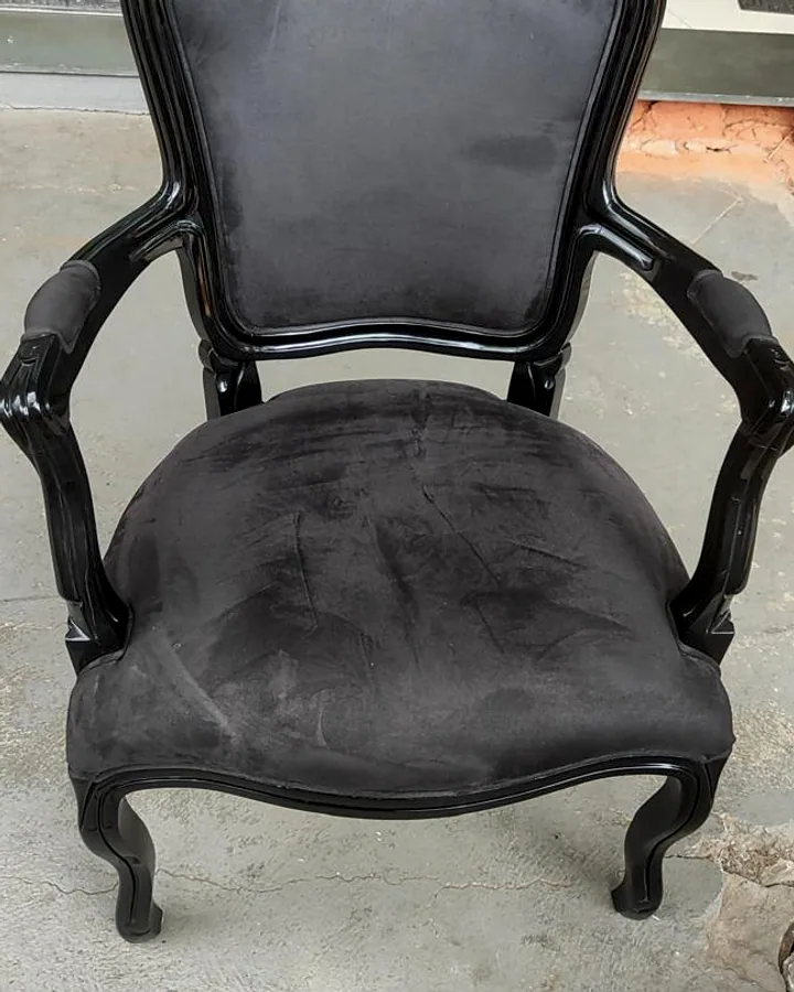 Poltrona clássica estilo Luís XV estofada em veludo preto com estrutura de madeira laqueada em preto sobre chão de cimento.