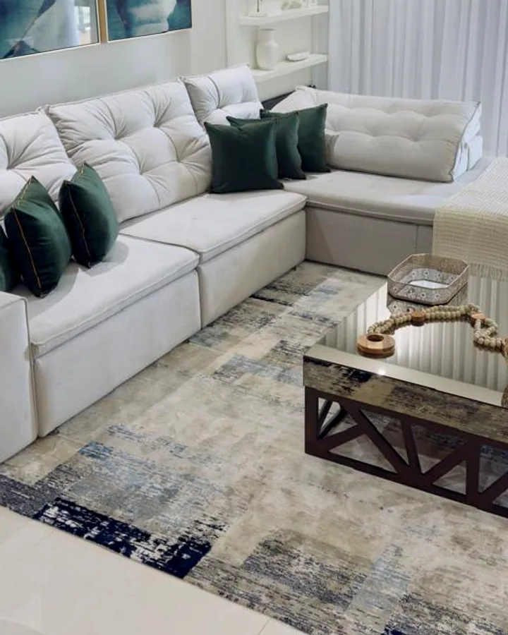 Amplo sofá claro com detalhes capitonados no assento e almofadas verdes, centralizado em uma sala de estar elegante e decorada.