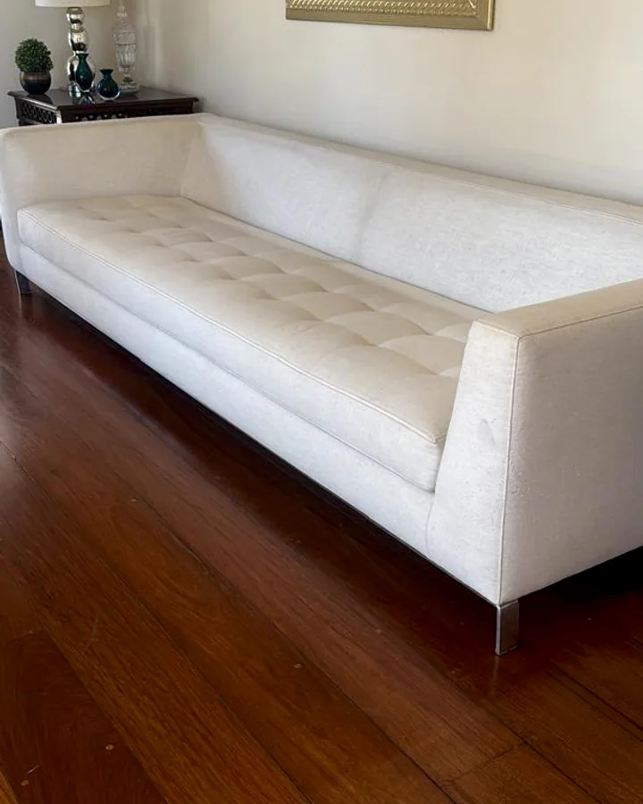 Sofá comprido com pés cromados e assento detalhado em capitonê, estofado em tecido claro sobre piso de madeira.