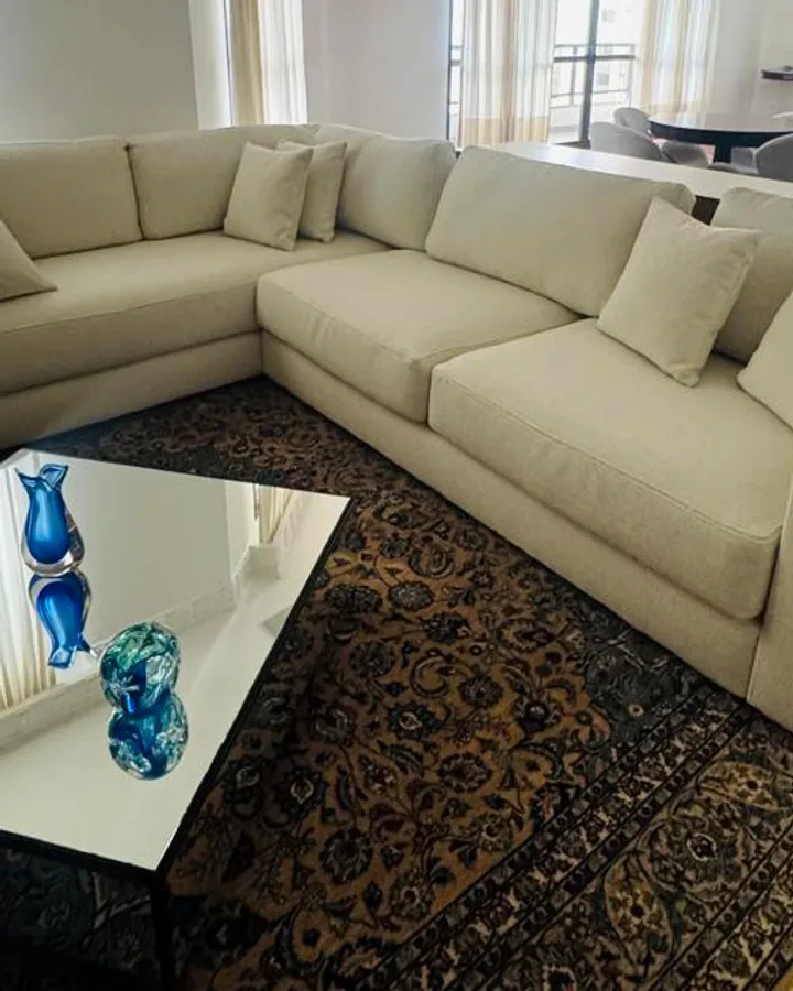 Sofá modular de canto em tecido claro em uma sala de estar ampla com mesa de centro espelhada e tapete clássico.