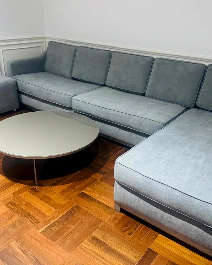 Sofá de canto espaçoso em tecido azul acinzentado com pufes e mesa de centro redonda em sala de piso de madeira.